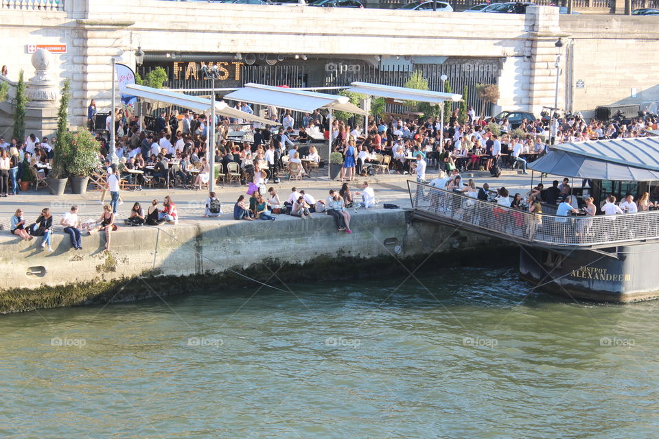 Some people are hanging out near the water while some of them sitting in the boat and ground restaurants in Paris.