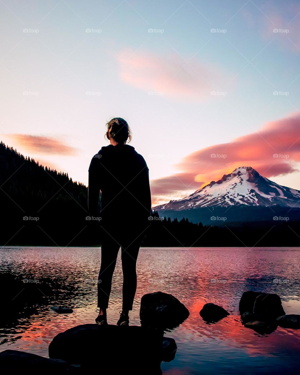 Woman stands at waters edge on stunning lake during colorful sunset over mountain 