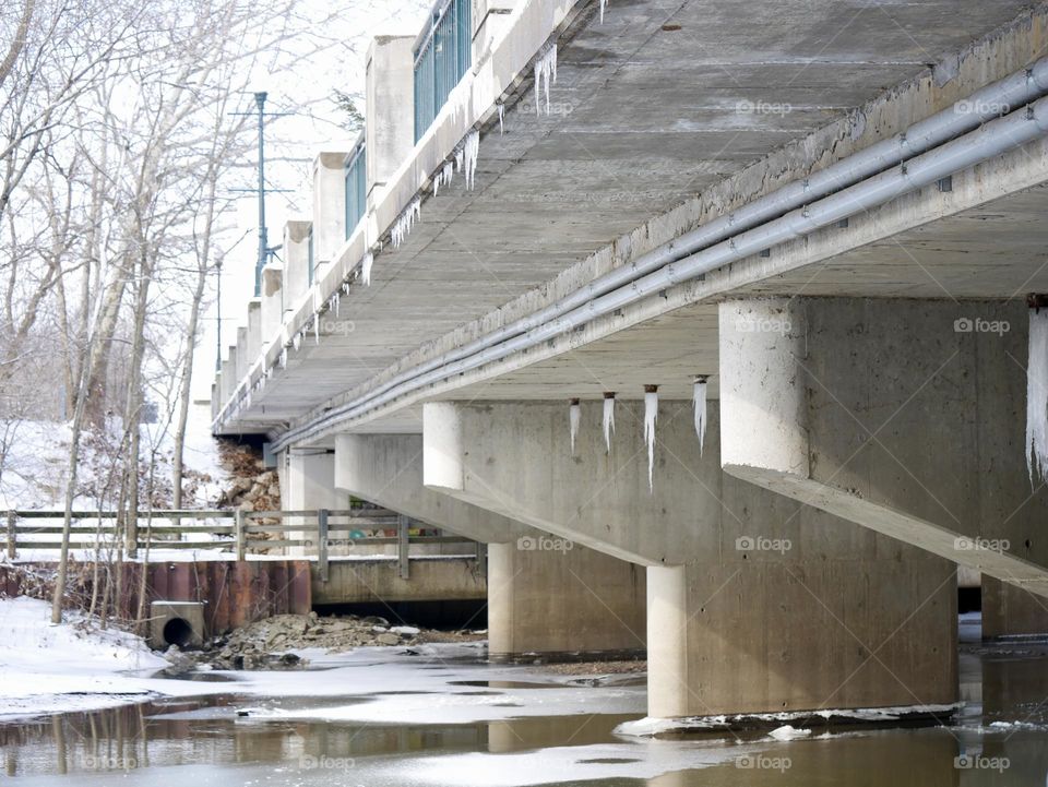 The underside of a city bridge, with ice hanging from the drains, and ice firming in the water underneath. 