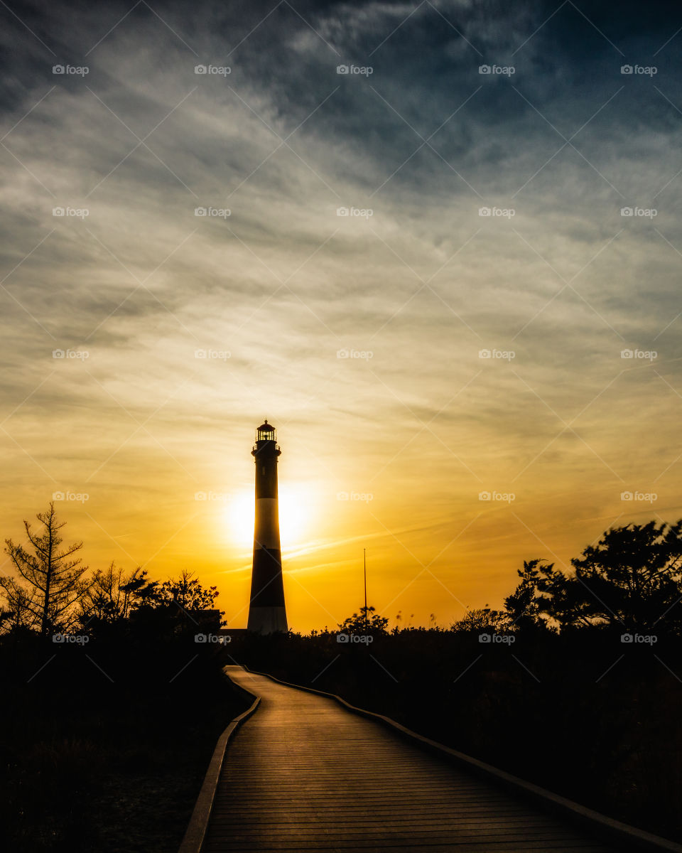 Silhouette of a wooden boardwalk leading to a lighthouse during a sunset. Orange warm tones from the sky reflecting light into the path