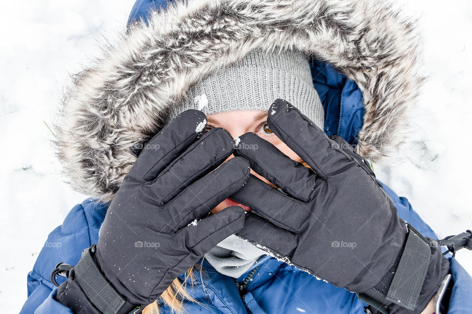 Close-up of a young millennial woman wearing a hooded winter coat in the snow and peeking through gloved hands with one eye