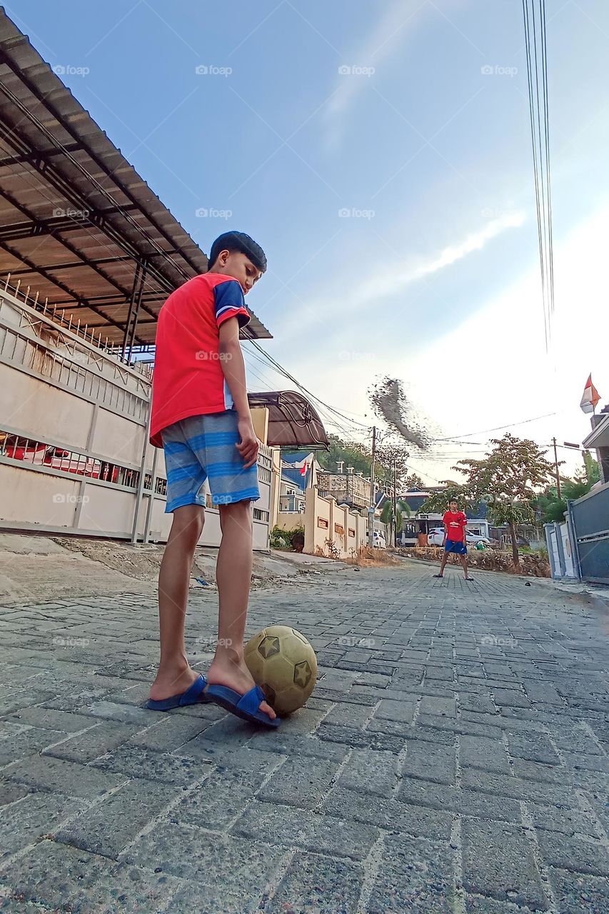 Bandar Lampung, Lampung, Indonesia - December 15, 2023: a child playing ball on a cobblestone street, with buildings and a clear sky in the background