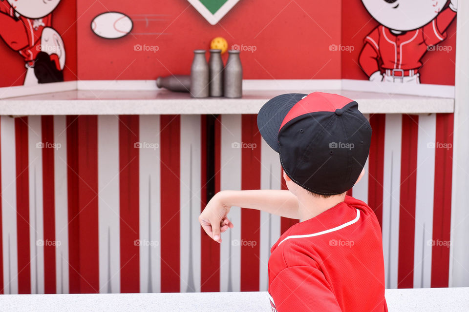 From behind candid shot of a young boy playing a ball toss carnival game outdoors in the summer