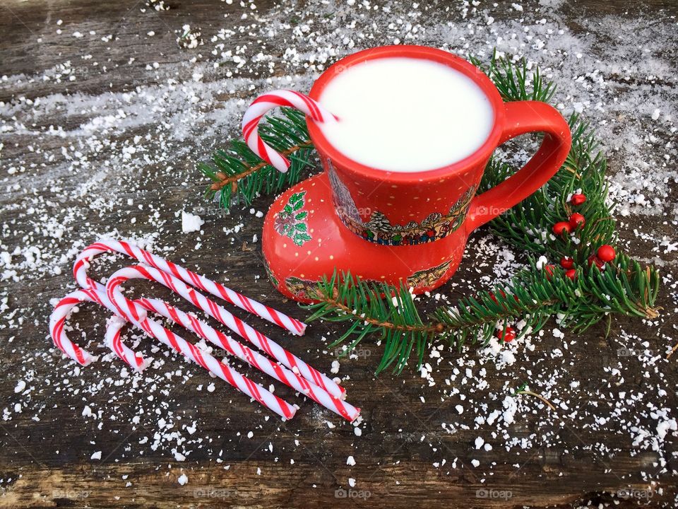 Boot shaped mug filled with milk and candy canes with candy canes and pine cone tree branches on snowy table 