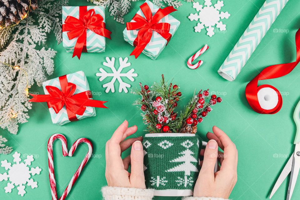 The hands of a young Caucasian girl hold a mug in a christmas sweater with fir branches, and around there are three gift boxes, decorative snowflakes, lollipops, wrapping paper, scissors, a ribbon, a fir branch and a garland on a green background, a