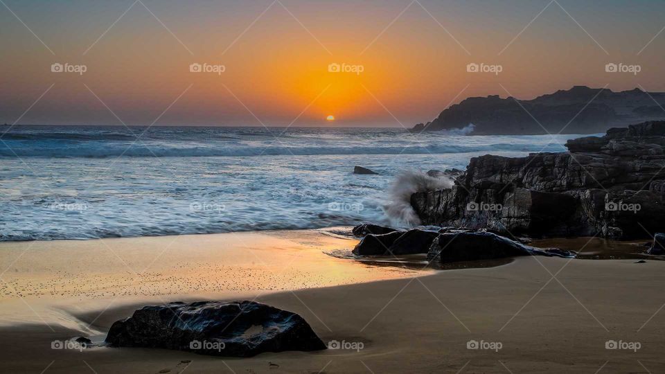 sunset on the coast the waves hitting the rocks and the sun submerging