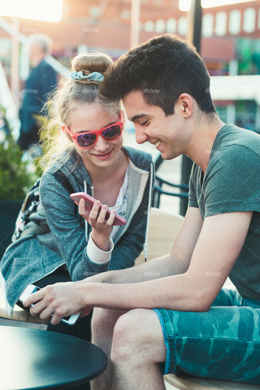 Couple of friends, teenage girl and boy, having fun using smartphones sitting in center of town, spending time together