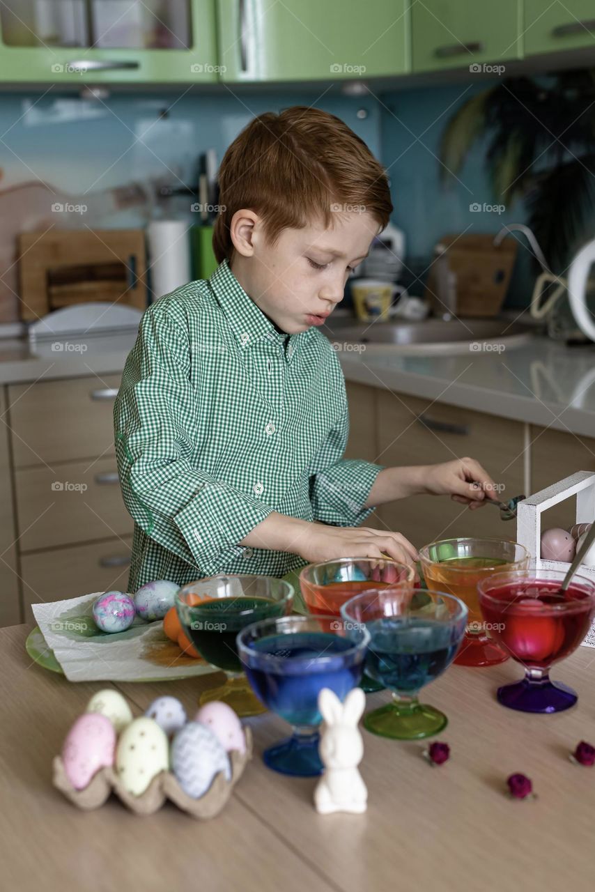 Red-haired boy paints eggs for Easter at home in the kitchen