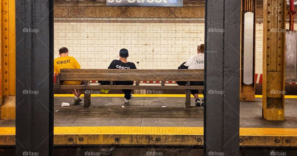 Three people with words printed on their T-shirts, sitting on a bench, waiting for a train in a New York City subway station 