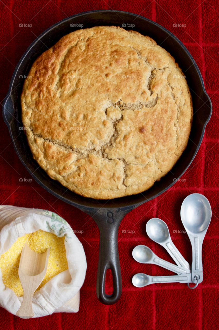 Flat lay of sourdough cornbread in a cast iron skillet, cornmeal and wooden scoop in a flour sack, vintage measuring spoons on a red kitchen towel