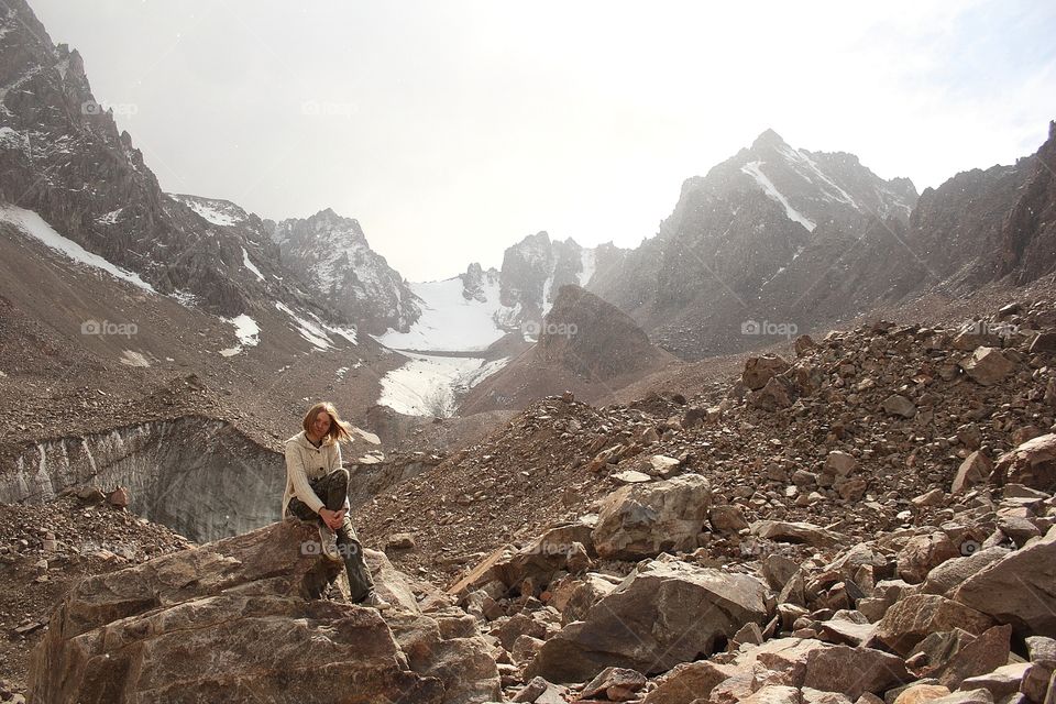 A girl in a knitted sweater with a hood sits on a rock under the falling snow in the mountains on the glacier