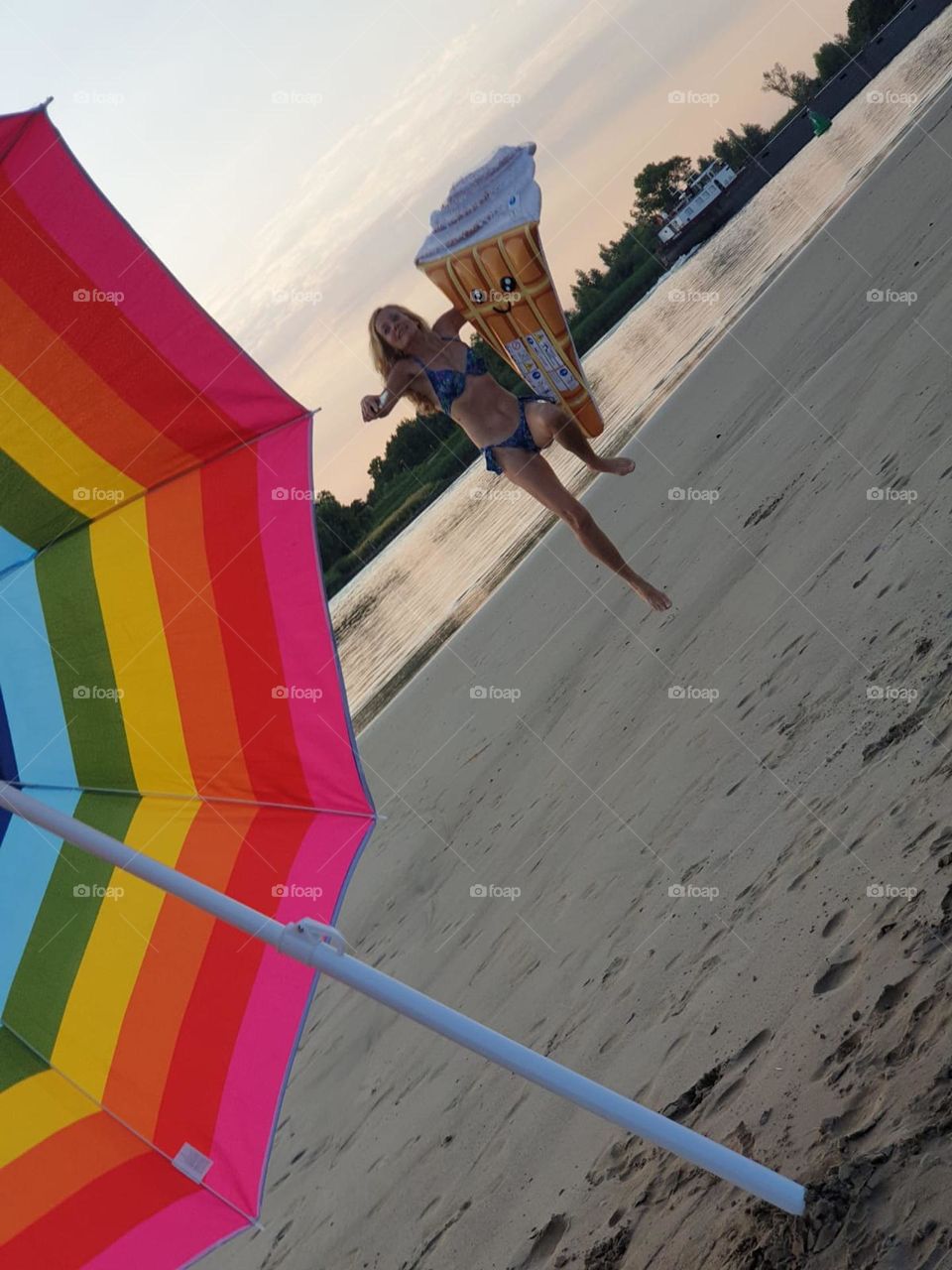 happy woman jumping on the sand of the beach holding an inflatable ice cream with a rainbow colored lgbt sun umbrella at the foreground