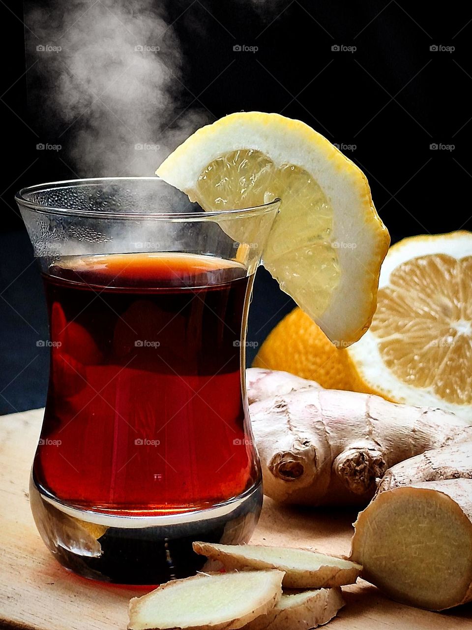 Still life of tea drinking.  A glass of "armuda" with strong hot tea from which steam comes.  A slice of lemon is hung on the rim of the glass.  Nearby lies a ginger root, sliced ​​​​pieces of ginger and lemons. The background is black