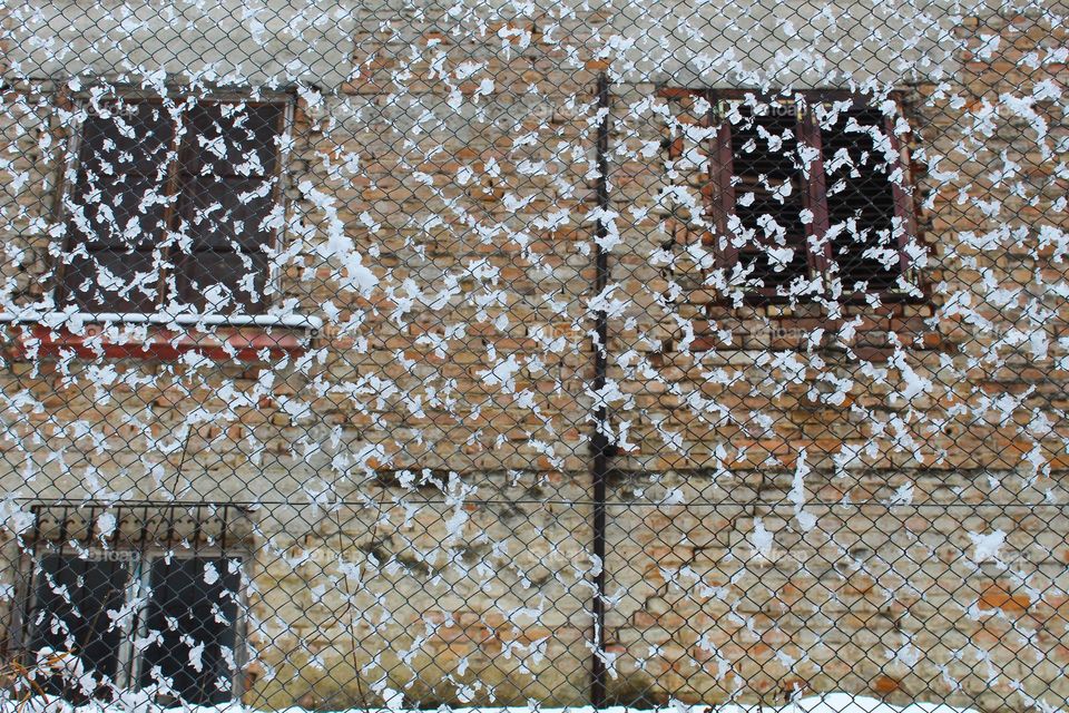 Snow on the wire fence in front of the abandoned and dilapidated red and white brick house