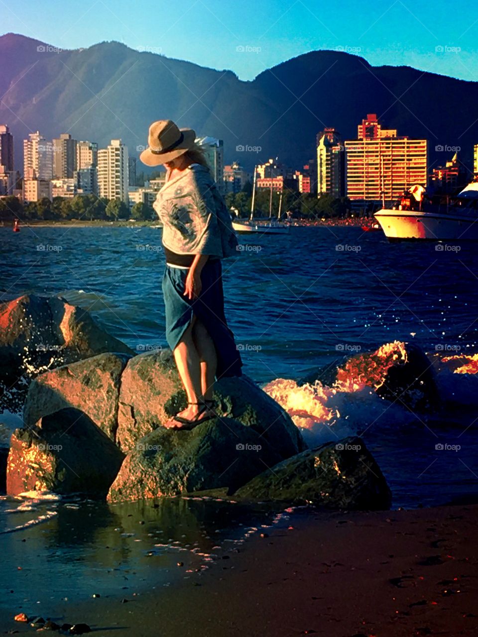 Woman in long skirt walking atop rocks by the ocean with view of Vancouver and sailboats 