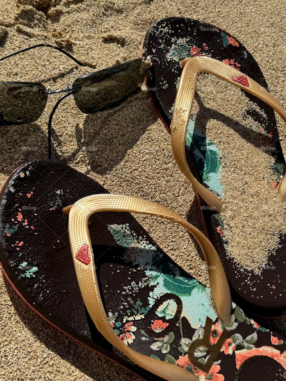 Worn Roxy brown floral pattern flip flops with gold straps and Maui Jim sunglasses in wet beach sand in Hawaii