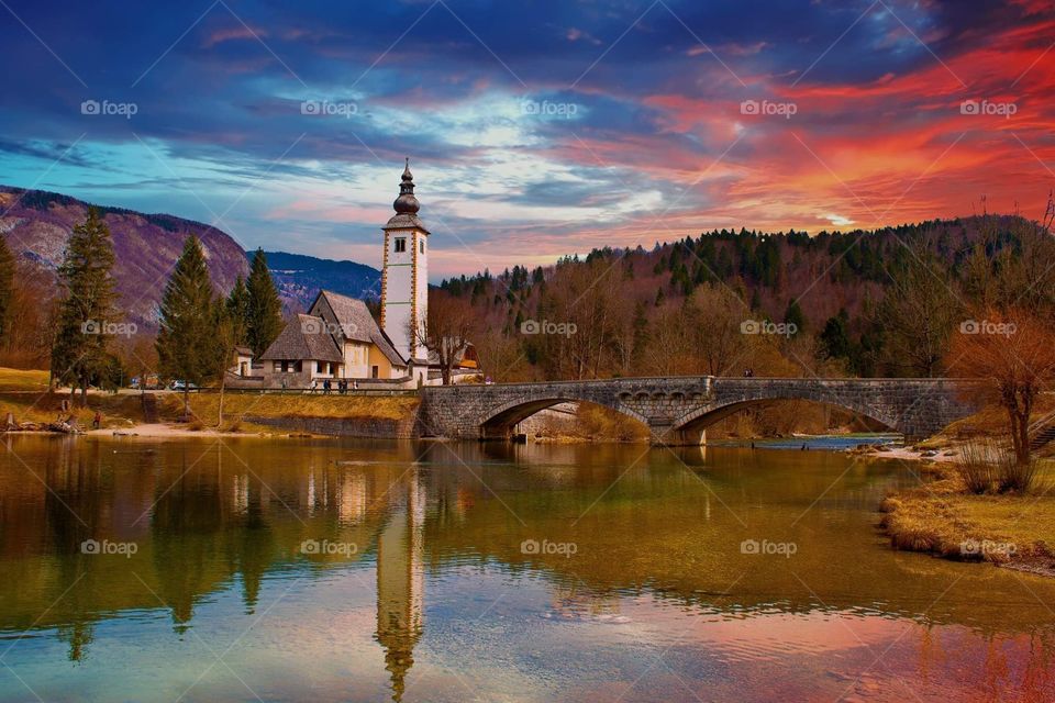 A red sunset sky at Lake Bohinj in Slovenia, with a bridge and chapel in the foreground, and their reflection in the lake.