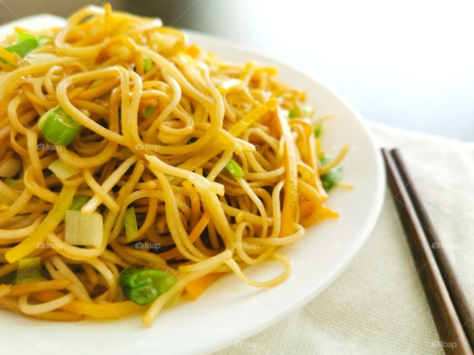 Chow mein with vegetables in a white plate with chopsticks on wood table.