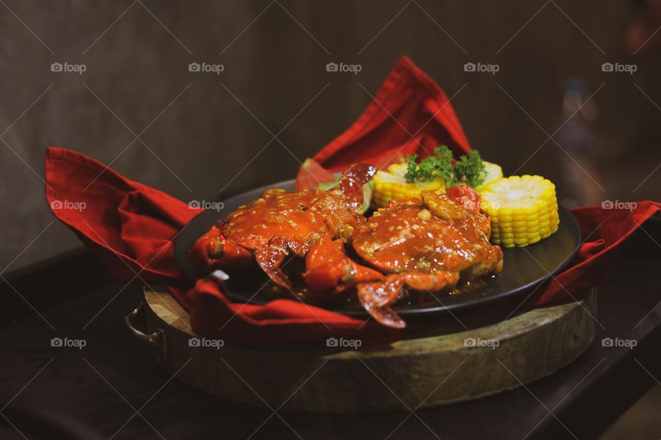 Crab flavored in creamy garlic sauce served with corn, tomatoes, lemon wedges and cherries on a black plate set on a red cloth set in a wooden setting. Asian flavor crab.