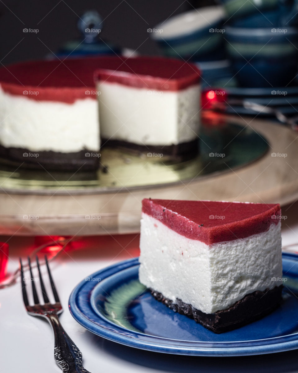 Piece of cheesecake with three colored layers is laying on blue plate. Red strawberry jelly on the top, Oreo layer on the bottom.