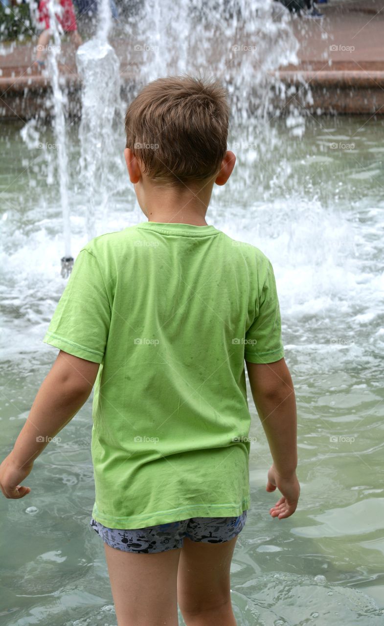 child boy in the water splash fountain, summer heat, city street view