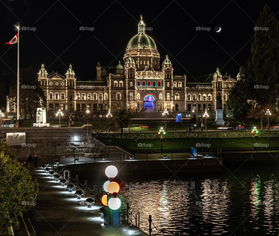 Historic government building lit up in Christmas lights and regular lights with moon above - the Legislature in Victoria, British Columbia, Canada