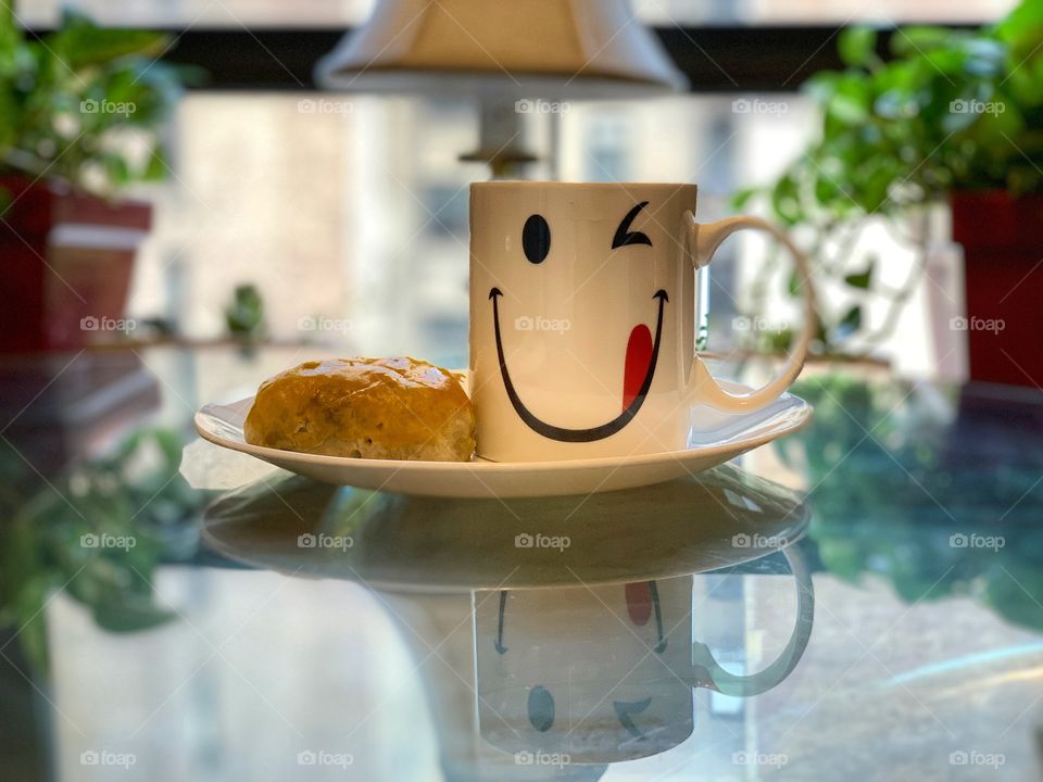 Reflection of a white cup with smiley design. Reflection of plate, leaves and lamp in a glass table.