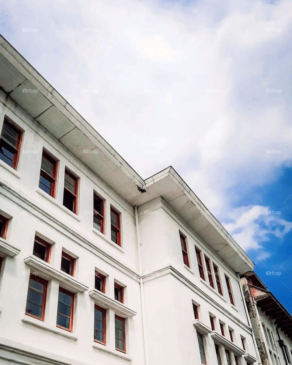 Part of a white building with brown framed windows, taken from a low angle with blue sky and white clouds in the background in low angle view