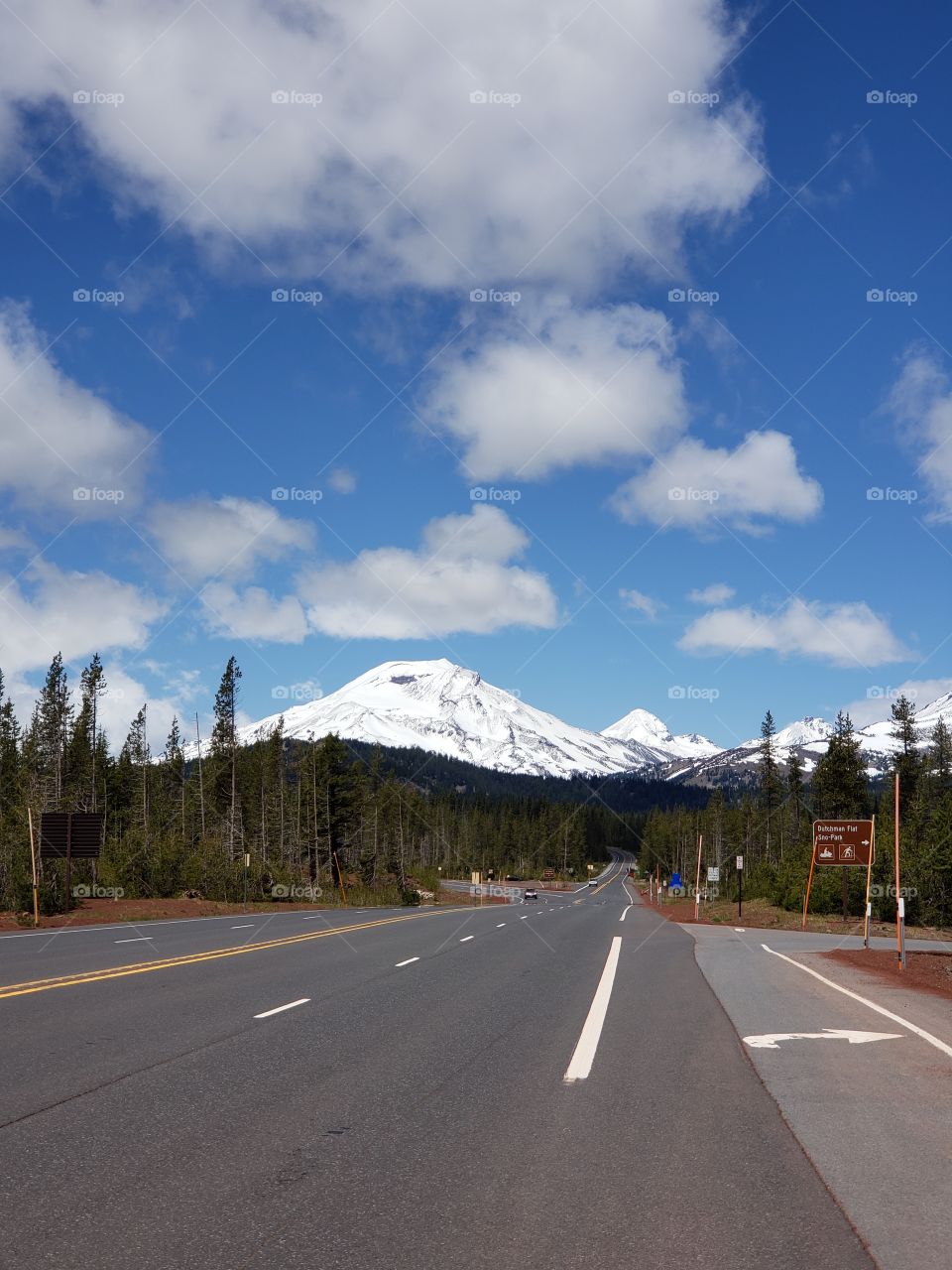 The snow covered South Sister in Oregon's Cascade Mountain Range as seen from Dutchman Flat looms large on the horizon of the Cascade Lakes Highway against a beautiful blue sky with fluffy white clouds on a sunny summer day in the forest.