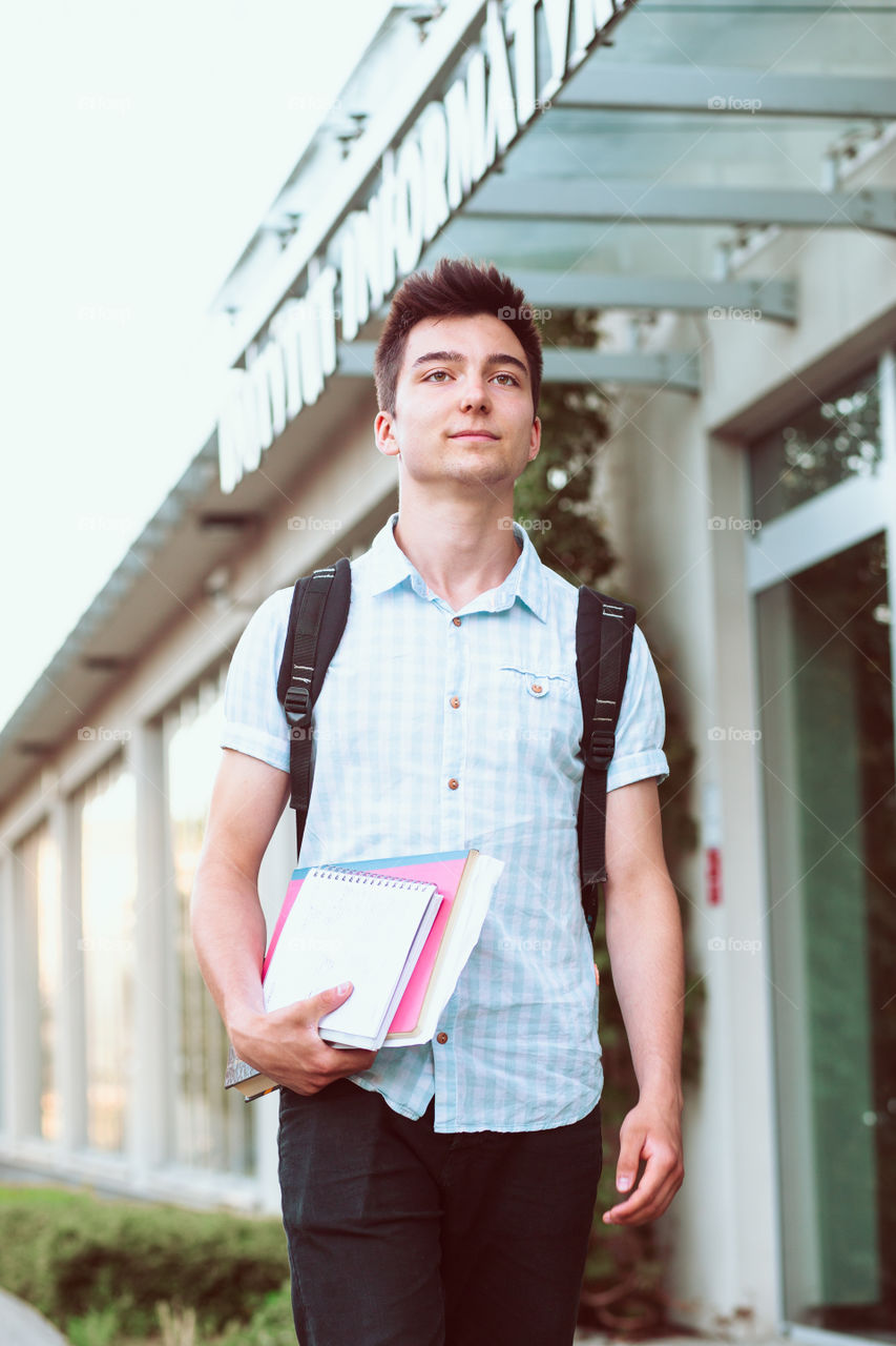 Student holding a notebook and carrying a backpack walking at the front of university building. Young smiling boy wearing blue shirt and dark jeans