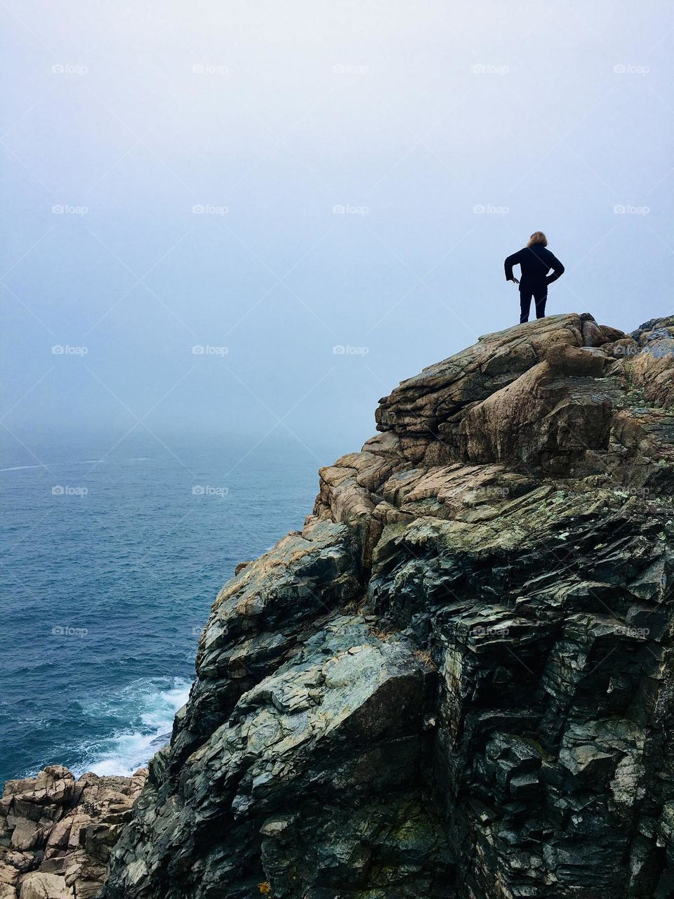 Woman overlooking the fog-covered ocean after hiking to the top of a rocky cliff during Spring in Maine.
