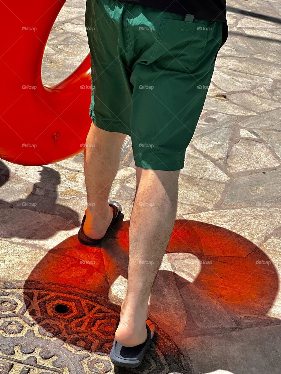 A man in green shorts is carrying a translucent red pool float and it casts a wonderful red shadow on the sidewalk