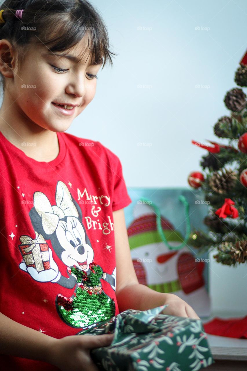 Happy girl in a bright t-shirt is opening her present by the Christmas tree