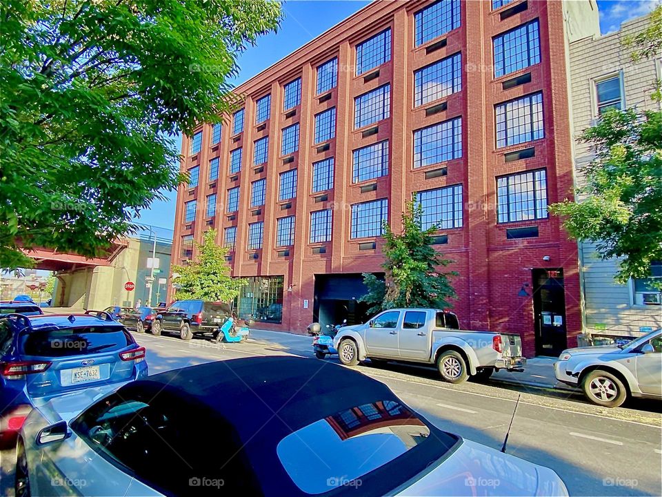 And from a little bit closer up we see the urban scenery in “Greenpoint”, Bklyn from before again. On the front right there is a white parked car with dark tinted windows. 2022. Hypnotic Productions