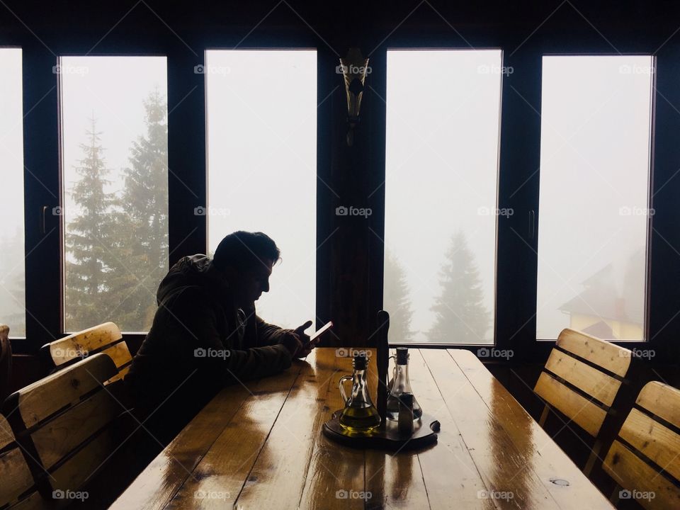 Single man in winter clothes texting at a table in a rustic restaurant with fog and trees seen out of the windows