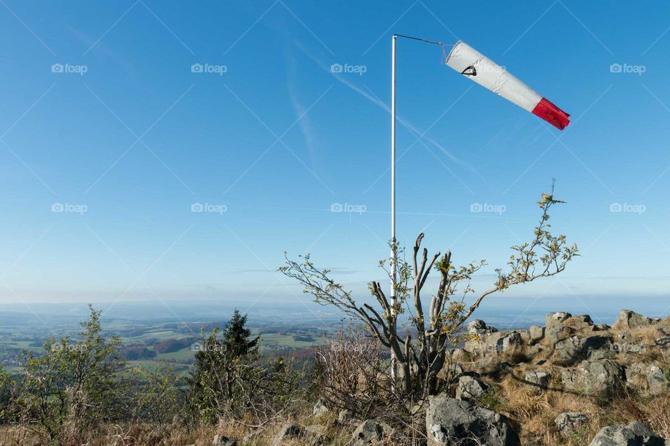 Windfahne auf einem Berg mit Seitenwind. Im Hintergrund ist der Blick ins Tal zu sehen. Im Vordergrund kleine Felsen und ein Gestrüpp. Zwei Tannen in mittlerer Entfernung. Aufgenommen an einem sonnigen Herbsttag bei wolkenlosem Himmel auf der Wasserkuppe in der Rhön.