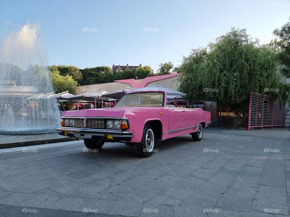 Classic pink Soviet cabriolet Chaika parked next to a fountain