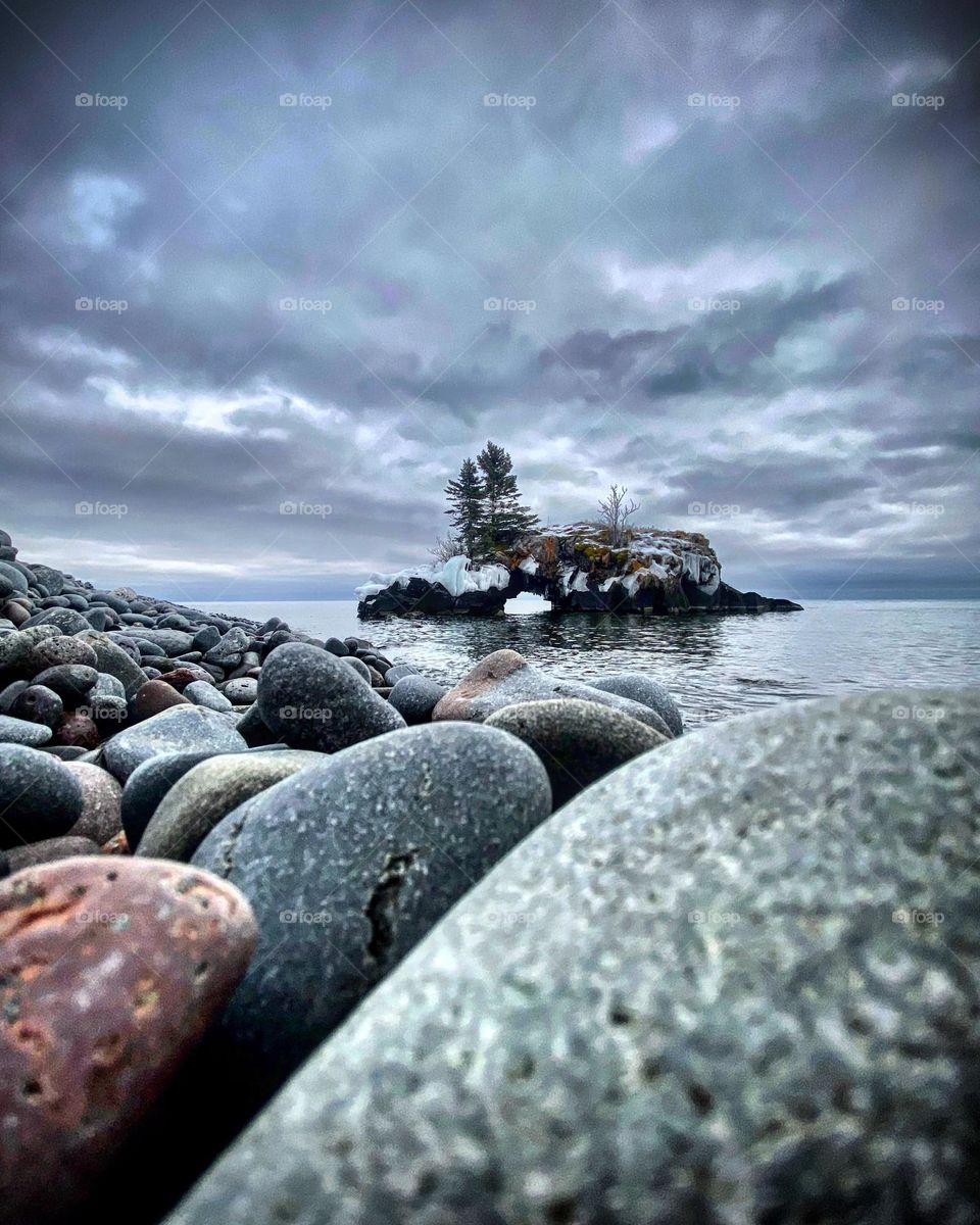 A moody winter day has the greys of the sky reflecting the greys of the rocks and lake as an arched island sits in the background 