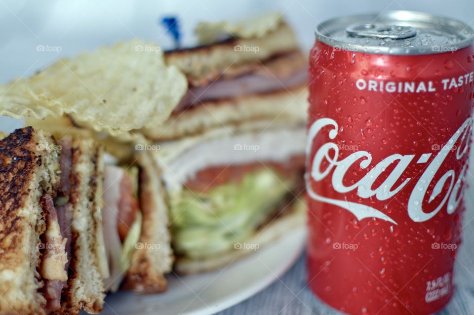 A can and a Bottle of Coca Cola with a club sandwich on a white rustic background 