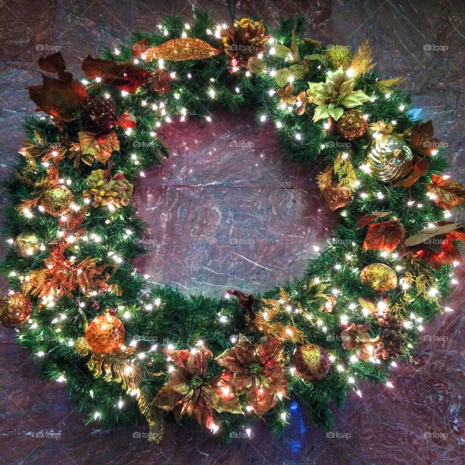 Large wreath with lights, pine cones, and greenery against a marble wall. 
