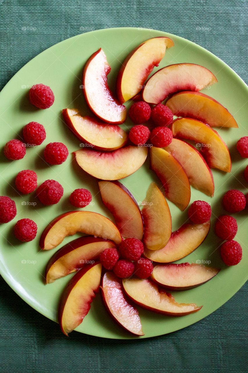 Flat lay of decorative arrangement of fresh nectarine slices and raspberries on a green plate with green background 