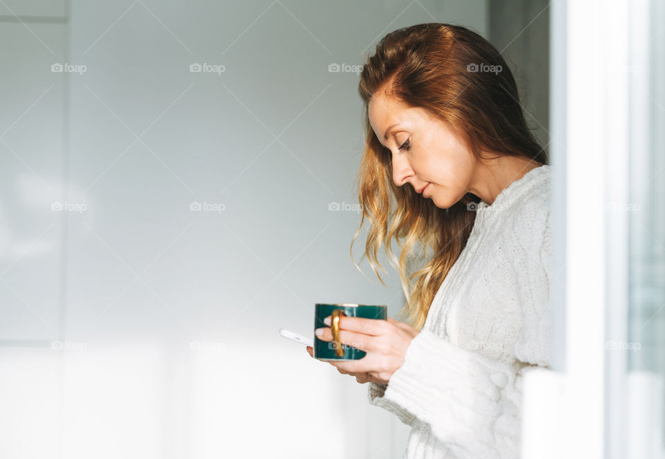 Young blonde woman with mobile and morning coffee or tea in cozy white knitted sweater in kitchen at home 