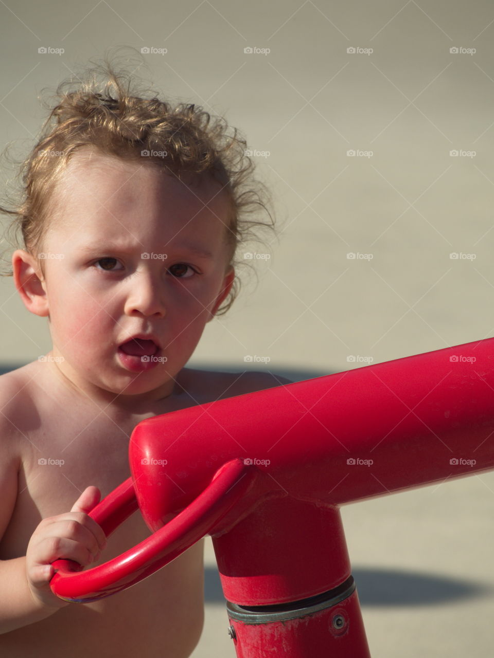 A toddler boy with curly hair plays with a water canon on a hot summer day. 