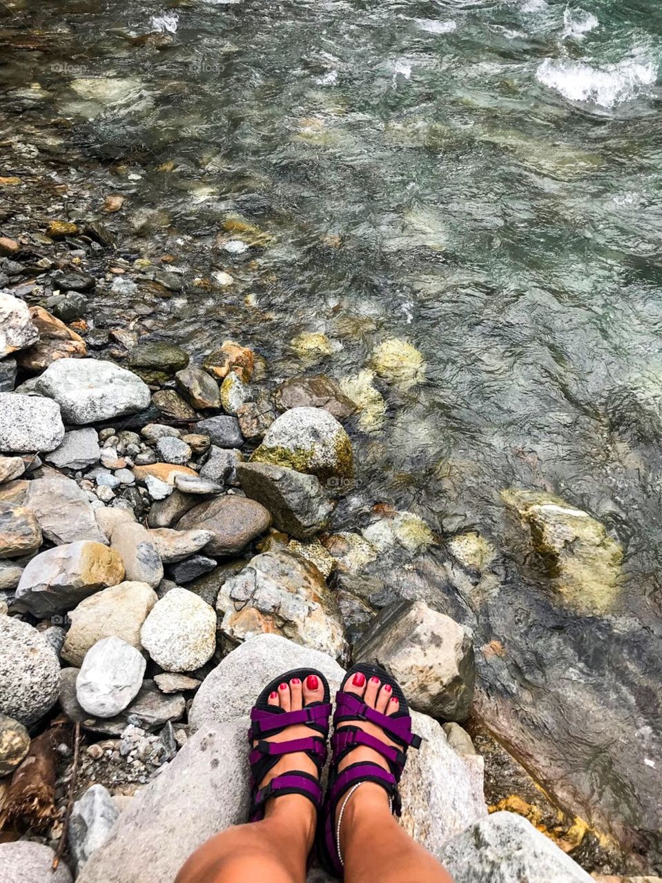 feet in purple shoes stand on rocks and water. The water is clear and cool