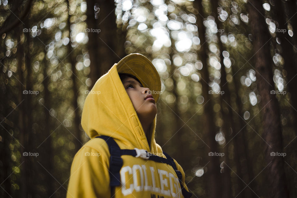 Seasonal outdoor close up portrait of a lonely Eurasian kid hiking in a pine forest wood.Natural setting, the boy is wearing yellow sweater with hood and cap, looking at his hand surrounded by pinetrees and magical day light bokeh