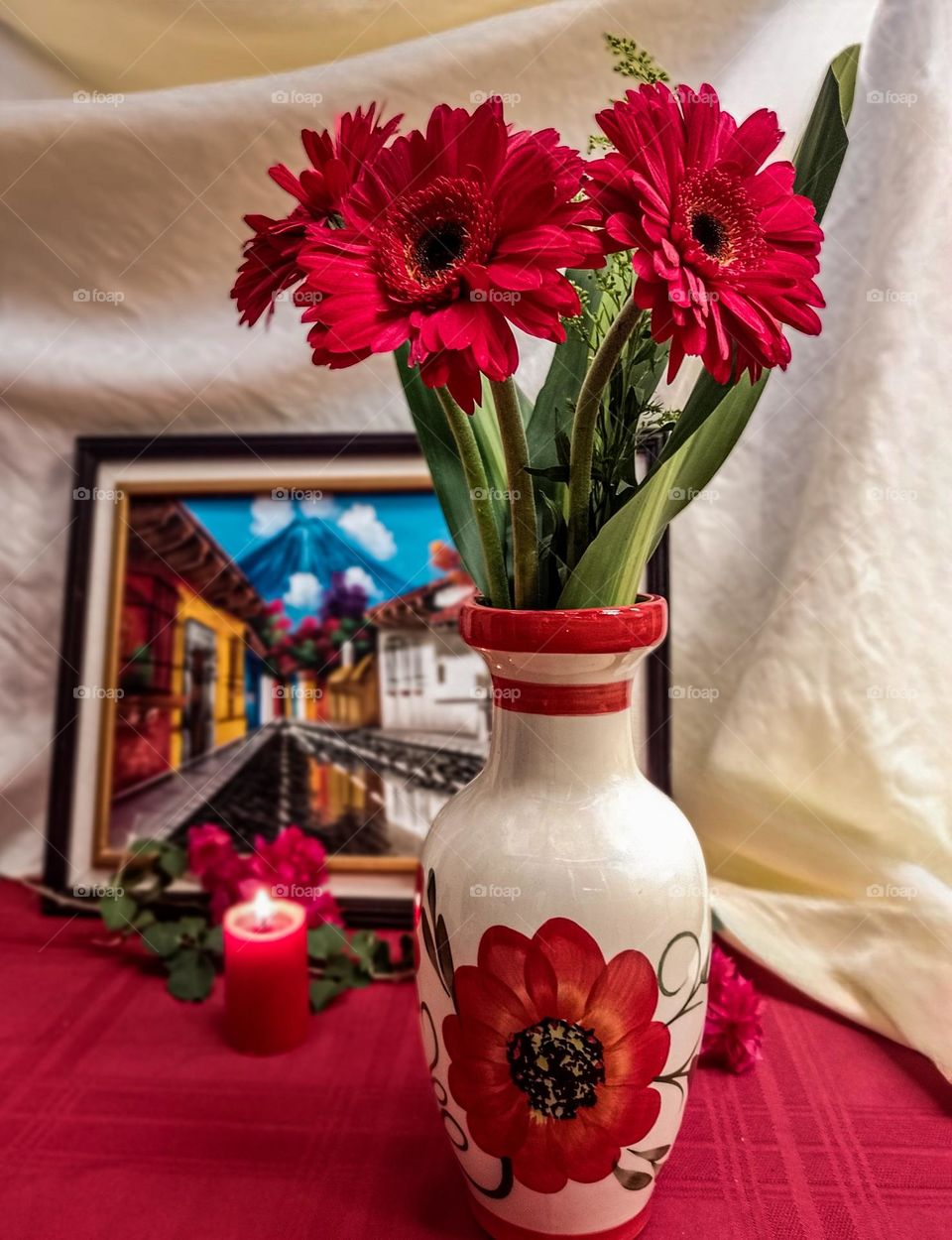 red flowers in a vase with a painting and a candle behind it, in portrait mode