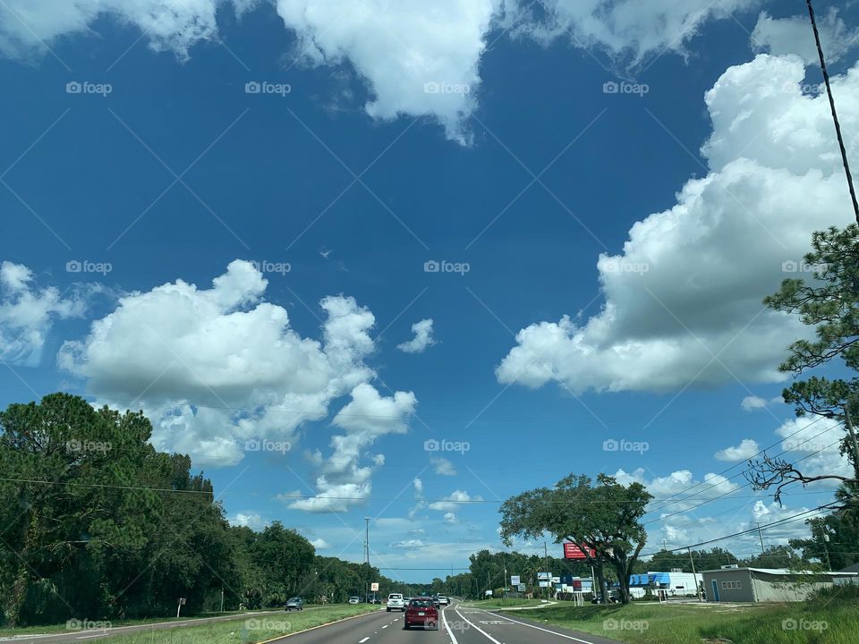 Commuting: vehicles and cars on the road traveling around with blue sky and grey cumulus around and green vegetation with trees and buildings.