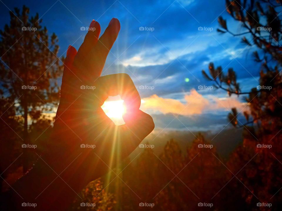 beautiful sunset in Nepal, enjoying with golden and warmth rays spreading from setting horizon hill, captured with hand and sun in between two fingers in a circular form, blue and cloudy sky, pine trees,