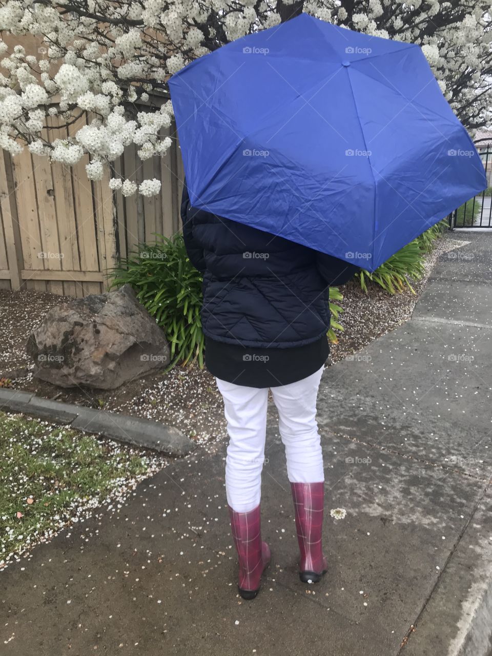 A girl walking in the rain holding a blue umbrella and wearing rain boots 
