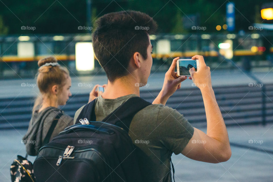 Young man with backpack taking photos using a smartphone in the city at night